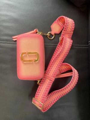 Rosa axelväska - Säljer en snygg rosa axelväska med guldiga detaljer och bred axelrem i mönstrad textil. Väskan har dubbla dragkedjor och en stilren logga framtill. Perfekt för dig som vill ha en färgklick till outfiten.