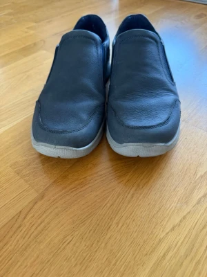Mörkblå loafers med vit sula - Snygga mörkblå loafers i skinn med elastiska partier på sidorna för enkel på- och avtagning. Skorna har en vit, sportig sula och en stilren design som passar till många olika outfits. Perfekta för dig som gillar en avslappnad men ändå trendig look.