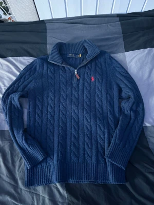Blå kabelstickad tröja Polo Ralph Lauren - Mörkblå kabelstickad tröja från Polo Ralph Lauren med halv dragkedja och hög krage. Tröjan har det klassiska röda Polo-logot på bröstet och ribbade muddar vid ärmslut och nederkant. Perfekt för dig som gillar en tidlös och snygg look.