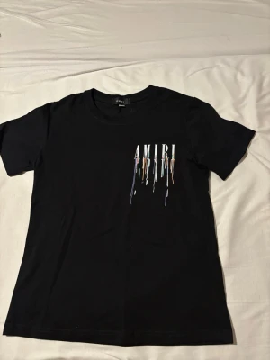 Svart AMIRI t-shirt med färgstänk - Svart t-shirt från AMIRI med tryckt logga och färgstänk-effekt både fram och bak. T-shirten har rund halsringning och korta ärmar. Perfekt för dig som gillar streetwear och vill sticka ut med en cool design.
