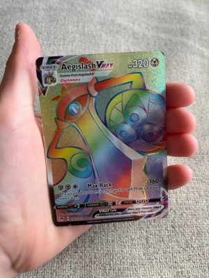 Aegislash VMAX Pokémonkort - Säljer ett Aegislash VMAX Pokémonkort med regnbågsglans och holografisk yta. Kortet har färgstarka detaljer och är en del av Sword & Shield-serien. Perfekt för samlare eller dig som vill boosta din lek med en VMAX-kort.