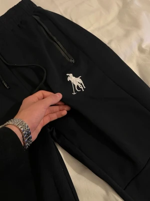 Svarta mjukisbyxor med logga - Svarta mjukisbyxor med dragsko i midjan och två fickor med dragkedja. På vänster ben finns en stor vit broderad logga med en häst och ryttare. Byxorna har en avslappnad passform och är gjorda i ett mjukt material.