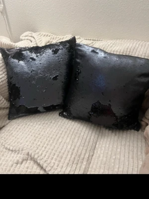 Svarta kuddar med paljetter - Två dekorativa kuddar med svarta paljetter som ger en skimrande och glansig effekt. Kuddarna är fyrkantiga och passar perfekt som snygg detalj i soffan eller på sängen. Materialet är textil med paljettöverdrag.