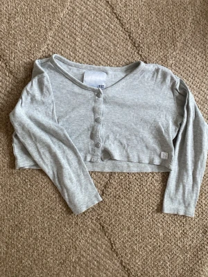 Ljusgrå kort cropped cardigan/top med knappar - Säljer en ljusgrå, ribbad cropped kort cardigan/top med långa ärmar och knäppning framtill. Tröjan har en enkel och stilren design som passar perfekt till jeans eller kjol. Materialet är mjukt och stretchigt för extra komfort.