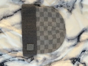 Grå mössa - Snygg gråmelerad mössa med rutmönster och bred ribbad uppvikt kant. Mössan har en liten patch framtill och är tillverkad i ett mjukt stickat material som håller värmen. Perfekt accessoar för kyliga dagar och enkel att matcha med olika outfits.