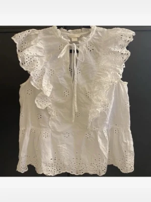 Vit broderad blus med volanger - Söt vit blus i bomull med broderade blommor och volangdetaljer. Blusen har knytband vid halsen och är ärmlös. Perfekt till jeans eller kjol för en trendig look.