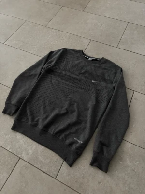 Mörkgrå sweatshirt från Nike - Mörkgrå sweatshirt från Nike med diskret svart mönster över bröstet och det klassiska Nike-loggan broderad på vänster sida. Tröjan har rund halsringning, långa ärmar och ribbade muddar vid ärmslut och nederkant. Perfekt för en sportig och avslappnad stil.