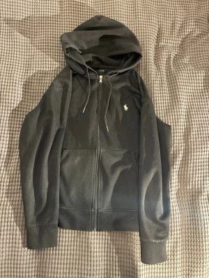 Ralph lauren hoodie - Säljer en svart hoodie med dragkedja och huva. Hoodien har två fickor framtill och justerbara snören i huvan. På bröstet finns en liten broderad logga i vitt. Perfekt för en chill och avslappnad stil. Tvättas innan den skickas.