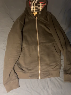 Svart hoodie med dragkedja och rutigt foder - Säljer en svart hoodie med dragkedja och klassiskt rutigt foder i huvan och på insidan. Hoodien har två stora fickor framtill och ribbade muddar vid ärmar och midja. Perfekt för dig som gillar stilrena detaljer och bekväma plagg.