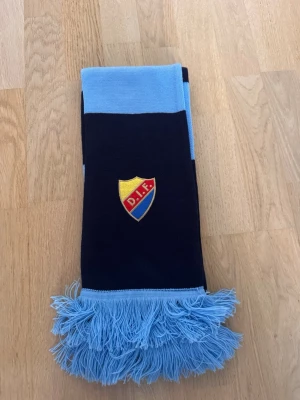Randig DIF halsduk i blått - Snygg halsduk med breda marinblå och ljusblå ränder samt DIF-emblem broderat på ena sidan. Halsduken har fransar i ljusblått i båda ändar och är perfekt för att visa ditt supporterskap. Materialet är mjukt och stickat, vilket gör den skön att ha på sig.