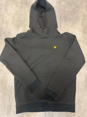 Svart hoodie från Lyle & Scott - Snygg svart hoodie från Lyle & Scott med klassisk huva och dragsko. På bröstet sitter den ikoniska gula loggan. Tillverkad i mjukt material som känns skönt mot huden. Perfekt för dig som vill ha en stilren och enkel look.