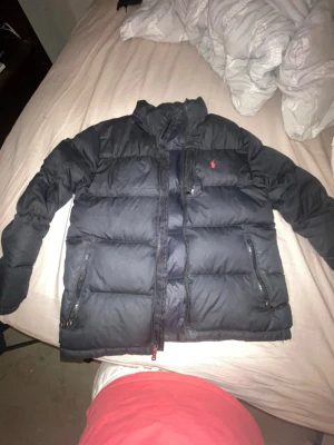 Svart pufferjacka från Polo Ralph Lauren - Säljer en svart pufferjacka från Polo Ralph Lauren med orange logga på bröstet. Jackan har hög krage, dragkedja framtill och två sidofickor med dragkedja samt en bröstficka. Perfekt för kalla vinterdagar och riktigt skön att bära.