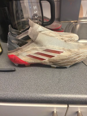 Adidas grå-röda fotbollsskor utan snörning - Säljer ett par Adidas fotbollsskor i grått och rött med tre röda ränder på sidan. Skorna har en modern, snörningsfri design och ovandel i mesh-liknande material för bra ventilation. Yttersulan är anpassad för spel på gräs. Perfekta för dig som vill ha stil och funktion på planen. Skruvdobbar