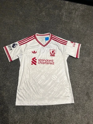 Liverpool borta 26/27 - Snygg vit Liverpool fotbollströja från Adidas med röda detaljer och klubbmärke på bröstet. Tröjan har röda ränder på axlarna, Premier League-märke på ärmen och sponsorlogga framtill. Tillverkad i lätt och ventilerande material, perfekt för match eller träning.