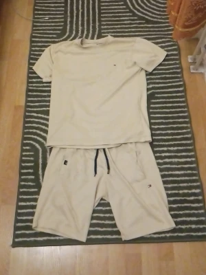 Beige loungewear-set från Tommy Hilfiger - Snyggt och bekvämt loungewear-set i beige från Tommy Hilfiger. Setet består av en t-shirt och shorts i mjukt, strukturerat material med diskret logga på både tröja och shorts. Perfekt för chill dagar hemma.