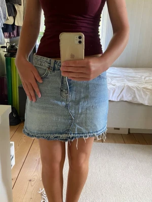 Blå jeanskjol med fransig kant - Snygg blå jeanskjol i kort modell med rå, fransig kant nertill. Kjolen har fyra fickor, bälteshällor och stängs med knapp och dragkedja framtill. Perfekt till sommaren och enkel att styla med olika toppar.