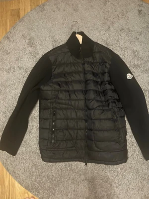 Svart pufferjacka från Moncler - Snygg svart pufferjacka från Moncler med quiltad front och släta ärmar. Jackan har hög krage, dragkedja framtill och två fickor med dragkedja. Moncler-logga på ena ärmen ger en exklusiv touch. Perfekt för kyliga dagar. Ända som är fel på jackan är att moncler märket i nacken har åkt bort annars inget fel på jackan.