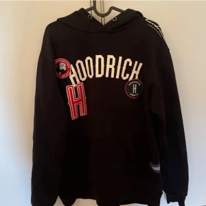 Svart hoodie med HOODRICH-tryck - Säljer en svart hoodie från Hoodrich med stora vita bokstäver på bröstet och flera patchar i rött och vitt. Hoodien har huva med dragsko och en klassisk känguruficka framtill. Perfekt för dig som gillar streetwear och vill sticka ut.