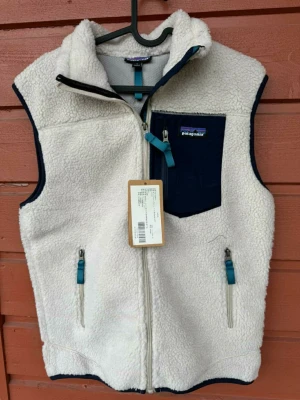 Patagonia väst - Mysig vit fleeceväst från Patagonia med marinblå detaljer och dragkedjor. Västen har en hög krage, två sidofickor med dragkedja och en bröstficka i marinblått tyg. Perfekt för lager-på-lager och outdoor-stil. Ej autentisk 