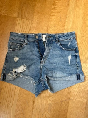 Blå slitna jeansshorts - Säljer ett par blå jeansshorts med slitna detaljer och fransig kant. Shortsen har klassisk femficksdesign, knappgylf och råa kanter för en avslappnad look. Perfekta för varma dagar och en chill stil.