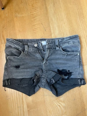 Grå slitna jeansshorts med detaljer - Säljer ett par grå jeansshorts med slitna detaljer och råa kanter. Shortsen har fem fickor, knappgylf och dekorativa sömmar på bakfickorna. Perfekta för en avslappnad och trendig look.