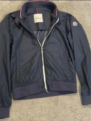 Mörkblå vindjacka från Moncler - Går inte ned i pris!  Den är helt äkta ny kr 10 00 kr har NFC tagg 👍 kan senda bild Snygg mörkblå vindjacka från Moncler med vit dragkedja, ribbade muddar och krage med röd och vit detalj. Jackan har Moncler-logga på ärmen och är tillverkad i ett lätt, glansigt material som står emot vind. Perfekt för dig som vill ha en stilren och sportig look.