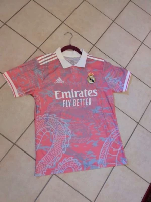 Rosa Real Madrid tröja Adidas - Säljer en rosa Real Madrid matchtröja från Adidas med vit krage och detaljer. Tröjan har ett drakmönster i ljusblått, klubbmärke på bröstet och 'Emirates Fly Better' tryckt framtill. Perfekt för dig som älskar fotboll och vill sticka ut på planen.