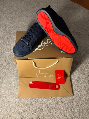 Mörkblå sneakers från Louboutin - Snygga mörkblå sneakers från Christian Louboutin i mocka med klassisk röd sula och ton-i-ton snörning. Skorna har en stilren och modern design med låg profil och exklusiva detaljer. Perfekta för dig som vill sticka ut med en ikonisk look.