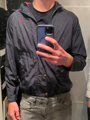 Polo Ralph lauren windbreaker  - Snygg marinblå vindjacka från Polo Ralph Lauren med röd luva och röd logga på bröstet. Jackan har dragkedja, tryckknappar och två fickor framtill. Perfekt för blåsiga dagar och enkel att matcha med olika outfits. Modellen är 178cm och väger 65kg