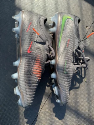 Nike svarta fotbollsskor med färgdetaljer - Säljer ett par svarta Nike fotbollsskor med coola detaljer i rött och grönt. Skorna har snörning och en modern, strömlinjeformad design med dobbar för bra grepp på planen. Perfekta för dig som vill sticka ut på fotbollsplanen!