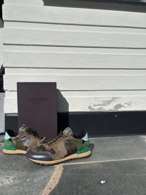 Valentino rockrunners  - Snygga sneakers från Valentino med kamouflagemönster i grönt, brunt och svart. Skorna har detaljer i mocka och läder, samt en cool grön och turkos häl. Yttersulan är beige med svarta nitar. Perfekta för dig som vill sticka ut med stilrena och unika sneakers.
