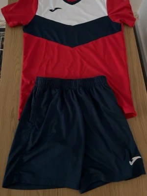 Röd och marinblå fotbollsset Joma - Säljer ett sportigt fotbollsset från Joma med röd och marinblå t-shirt samt matchande marinblå shorts. Tröjan har vit och marinblå detalj upptill och korta ärmar. Materialet är lätt och andas, perfekt för träning eller match.