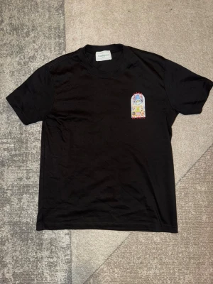 Svart t-shirt - Säljer en svart t-shirt med färgglatt tryck på bröstet och stort grafiskt motiv på ryggen. T-shirten har rund hals och korta ärmar. Perfekt för dig som gillar streetwear och unika prints.
