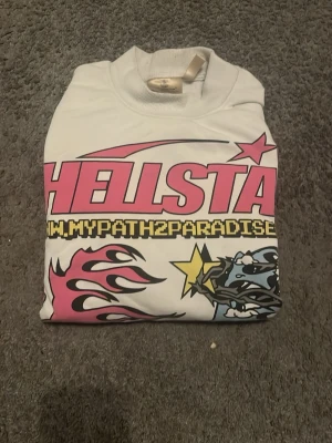 Vit Hellstar sweatshirt med tryck - Säljer en vit sweatshirt från Hellstar med stora grafiska tryck i rosa, svart, gult och blått både fram och bak. Tröjan har coola flames, stjärnor och texten 'MYPATH2PARADISE'. Långärmad och i mjukt material, perfekt för en streetwear-look. Köptes från USA Nwy för cirka 3000