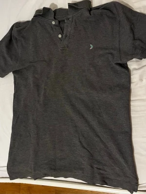 Mörk grå pikétröja från Boomerang - Snygg mörkgrå pikétröja i bomull med klassisk krage och tre knappar framtill. Tröjan har korta ärmar och en liten broderad detalj på bröstet. Perfekt till jeans eller shorts för en clean look.