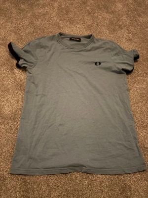 Blå t-shirt från Fred Perry - Snygg blå t-shirt från Fred Perry med klassisk logga broderad på bröstet. T-shirten har rund hals och korta ärmar, tillverkad i mjuk bomull för skön känsla. Perfekt till jeans eller shorts för en clean och enkel stil. Pris kan sänkas lite