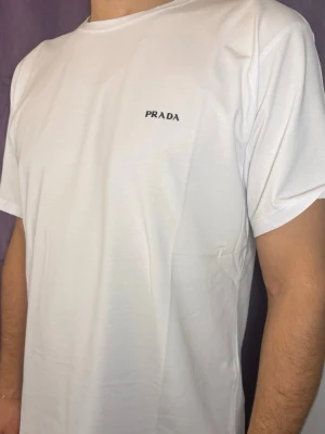 Vit t-shirt från Prada med logga - Vit t-shirt från Prada med diskret svart logga på bröstet. Klassisk rund hals och korta ärmar. T-shirten har en clean och enkel design som passar perfekt till jeans eller shorts. Materialet känns mjukt och bekvämt mot huden.