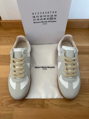 Maison Margiela sneakers - Snygga Maison Margiela sneakers i vitt och ljusgrått med beige skosnören och gummisula. Ovandelen är i en mix av mocka och läder för en clean och stilren look. Perfekta för dig som gillar minimalistisk design och streetwear-vibe.