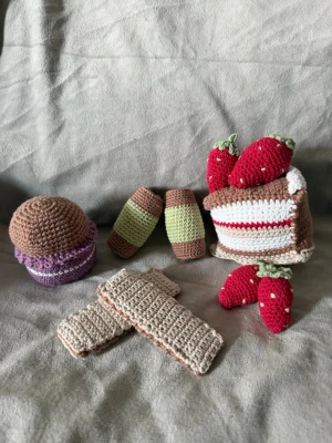 Virkade bakverk - Ett set med handgjorda virkade bakverk och jordgubbar. Innehåller en tårtbit med röda jordgubbar, två dammsugare, två wafers kex, en muffin samt två röda jordgubbar. Alla delar är virkade i bomullsgarn och har fina detaljer. Måtten är, Dammsugare 7x4cm, Muffin är 8x7cm, Jordgubbe stor är 5,5x3cm, Jordgubbe liten är 5x3cm, Tårtbit är 13x10cm, Wafers kex är 10,5x3,5cm