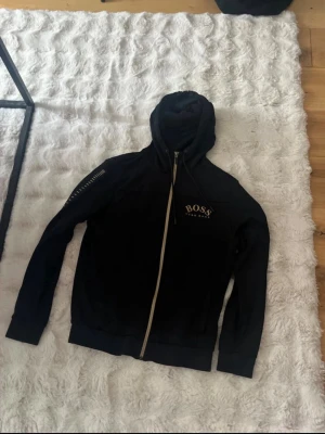 Svart hoodie med dragkedja från BOSS i S - Snygg svart hoodie från BOSS med dragkedja framtill och huva. Diskret BOSS-logga på bröstet och detaljer på ena ärmen. Tillverkad i mjukt material som känns skönt mot huden. Perfekt för en avslappnad och stilren look. Skriv vid minst fundering väldigt bra skick