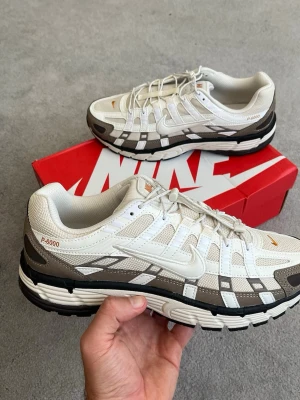 Nike P-6000 vita och bruna sneakers - Nike P-6000 sneakers i vitt och brunt med mesh och syntetiska överdrag. Skorna har en chunky sula och tydliga detaljer i orange. Swoosh-logga på sidan och P-6000-tryck på hälen. Perfekta för dig som gillar retroinspirerad stil.