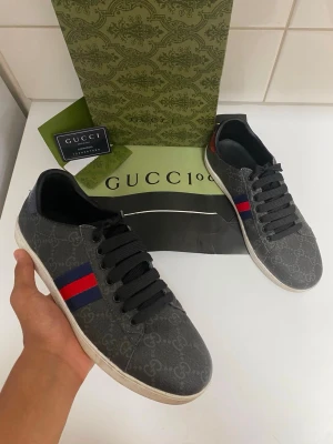 Gucci Skor - Snygga Gucci sneakers i mörkgrått med GG-monogram över hela skon. Klassiska röda och blå ränder på sidan och svarta skosnören. Yttermaterial i canvas och vit sula. Perfekta för dig som vill ha en stilren och exklusiv look.