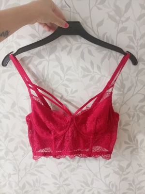 Röd spetsbralette med dubbla band - Snygg röd bralette i spets med dubbla axelband och dekorativa band framtill. Bh:n har hakknäppning bak och justerbara axelband. Spetsdetaljer längs nederkanten ger en extra feminin touch. Perfekt för dig som gillar trendiga underkläder. Nån tråd ä lite lös men funkar ändå 