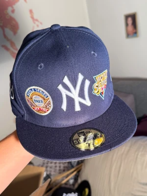 Mörkblå New York Yankees keps - Snygg mörkblå New York Yankees keps från New Era med broderade World Series-patchar och stor vit Yankees-logga framtill. Kepsen har platt skärm och detaljerad brodyr på sidan med texten 'World Series'. Perfekt för dig som gillar streetwear och sportig stil.