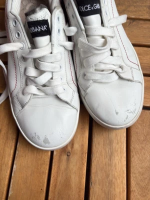 Vita Dolce & Gabbana sneakers - Snygga vita sneakers från Dolce & Gabbana med silvriga detaljer och svart logga på hälen. Skorna har vita snören, röda sömmar på sidan och är tillverkade i läder. Perfekta för dig som vill ha en clean och trendig look.