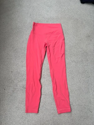 Korall lululemon leggings - Snygga rosa leggings med hög midja och långa ben. Tillverkade i ett stretchigt material som sitter skönt och följer kroppen. Perfekta för dig som vill ha en färgklick i garderoben och gillar bekväma plagg.