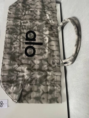 ALO totebag - Snygg shoppingväska från Alo i grått och beige med coolt batikmönster och svart logga på framsidan. Väskan har dubbla handtag och är tillverkad i slitstarkt tyg, perfekt för att bära med sig allt man behöver.