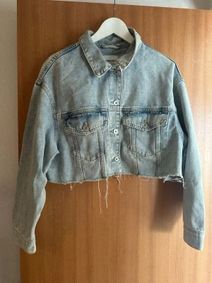 Cropped ljusblå jeansjacka - Cropped jeansjacka i ljusblå denim med rå, fransig nederkant. Klassisk krage, två bröstfickor med knappar och långa ärmar. Perfekt för dig som gillar en avslappnad och edgy stil.