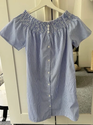 Blårandig kort klänning med knappar - Söt kort klänning med blå och vita ränder, smockad offshoulder-ringning och knappar framtill. Tillverkad i lätt bomull, perfekt för varma dagar. Kortärmad och avslappnad passform som ger en chill vibe.