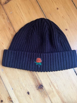 Svart ribbstickad mössa med hjortronmärke - Säljer en svart ribbstickad mössa med uppvikt kant och ett broderat märke av ett orange hjortron med gröna blad framtill. Mössan är i tjockt, mjukt material och har en klassisk, enkel form som passar till det mesta.